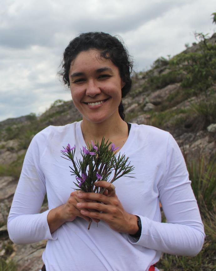 Jenifer de Carvalho Lopes, a woman stands holding a bundle of Vellozia with pink flowers, smiling